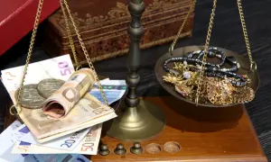 Waga szalkowa z euro i biżuterią. Na jednej szalce banknoty euro i monety, na drugiej złota biżuteria. Cena skupu 20 dolarów w złocie.