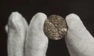 Dłonie w białych rękawiczkach trzymają starożytny, srebrny denar, jeden z najstarszych polskich monet.