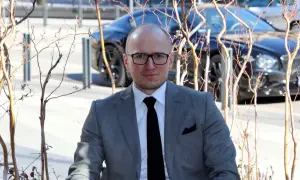 Mężczyzna w garniturze, z okularami, na tle luksusowego samochodu. Jego styl sugeruje sukces i przemyślane inwestycje rzeczowe.