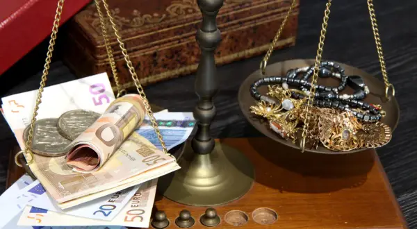 Waga szalkowa z euro i biżuterią. Na jednej szalce banknoty euro i monety, na drugiej złota biżuteria. Cena skupu 20 dolarów w złocie.